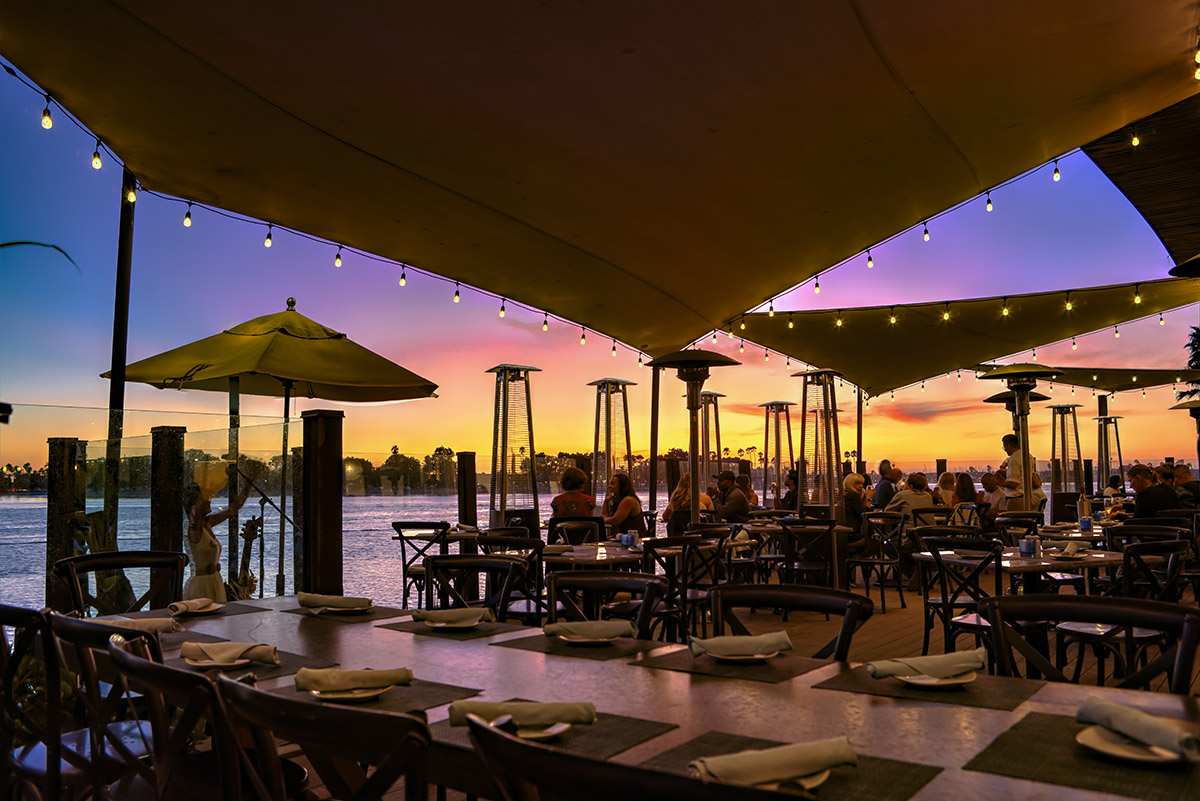 Outdoor waterfront dining at Tidal restaurant during sunset at Paradise Point Resort & Spa on Mission Bay, with string lights and guests enjoying the view.