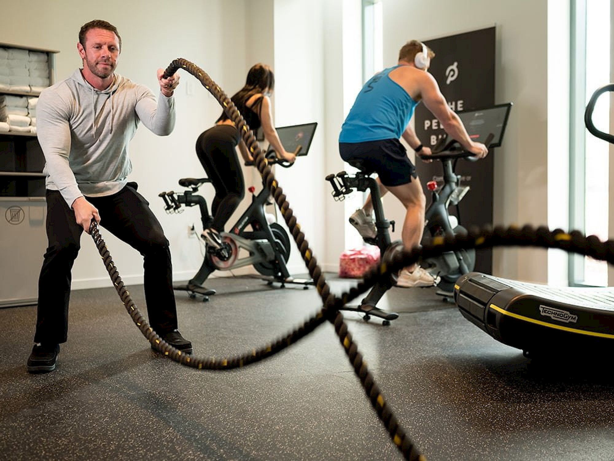 Guests exercising in the modern fitness center at Hotel ELEO at the University of Florida, featuring Peloton bikes, battle ropes, and Technogym workout equipment.