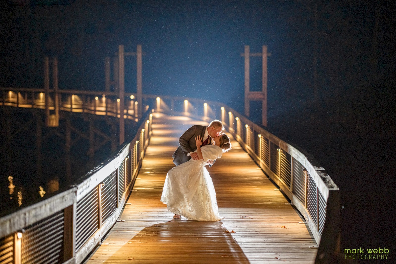 Bride and groom share a romantic kiss on a beautifully lit lakeside boardwalk at Stonewall Resort in Roanoke, West Virginia, during a nighttime wedding.